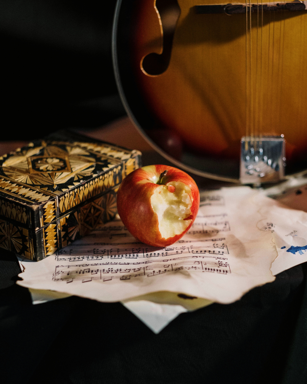 A bitten red apple on sheet music beside a decorative box and a guitar