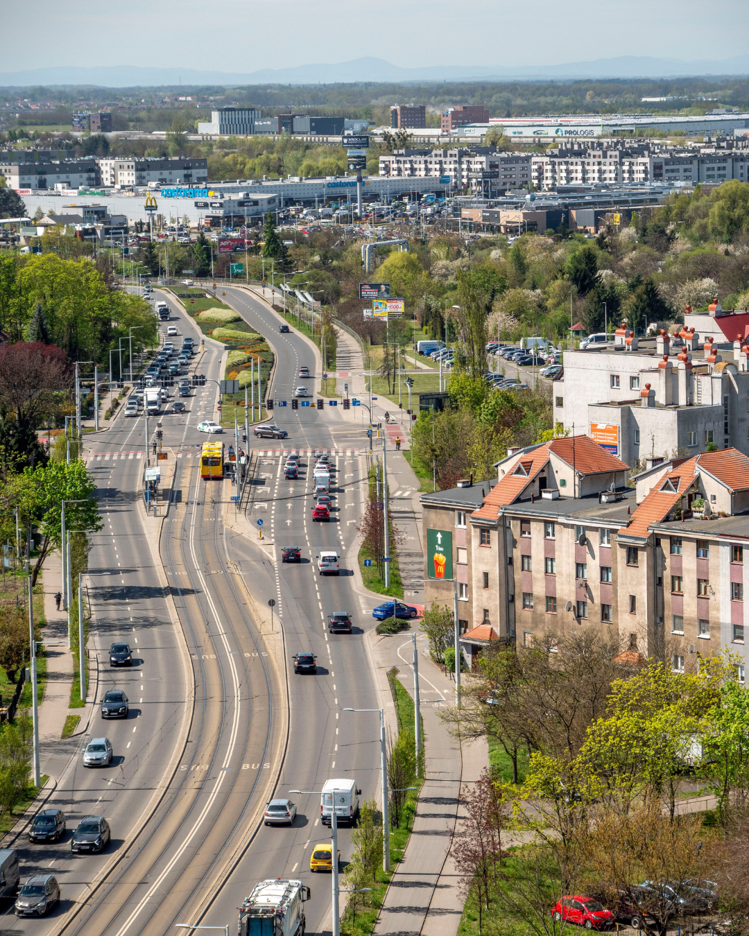 An elevated view of a broad city avenue, tram tracks, trees, and a distant horizon with low hills