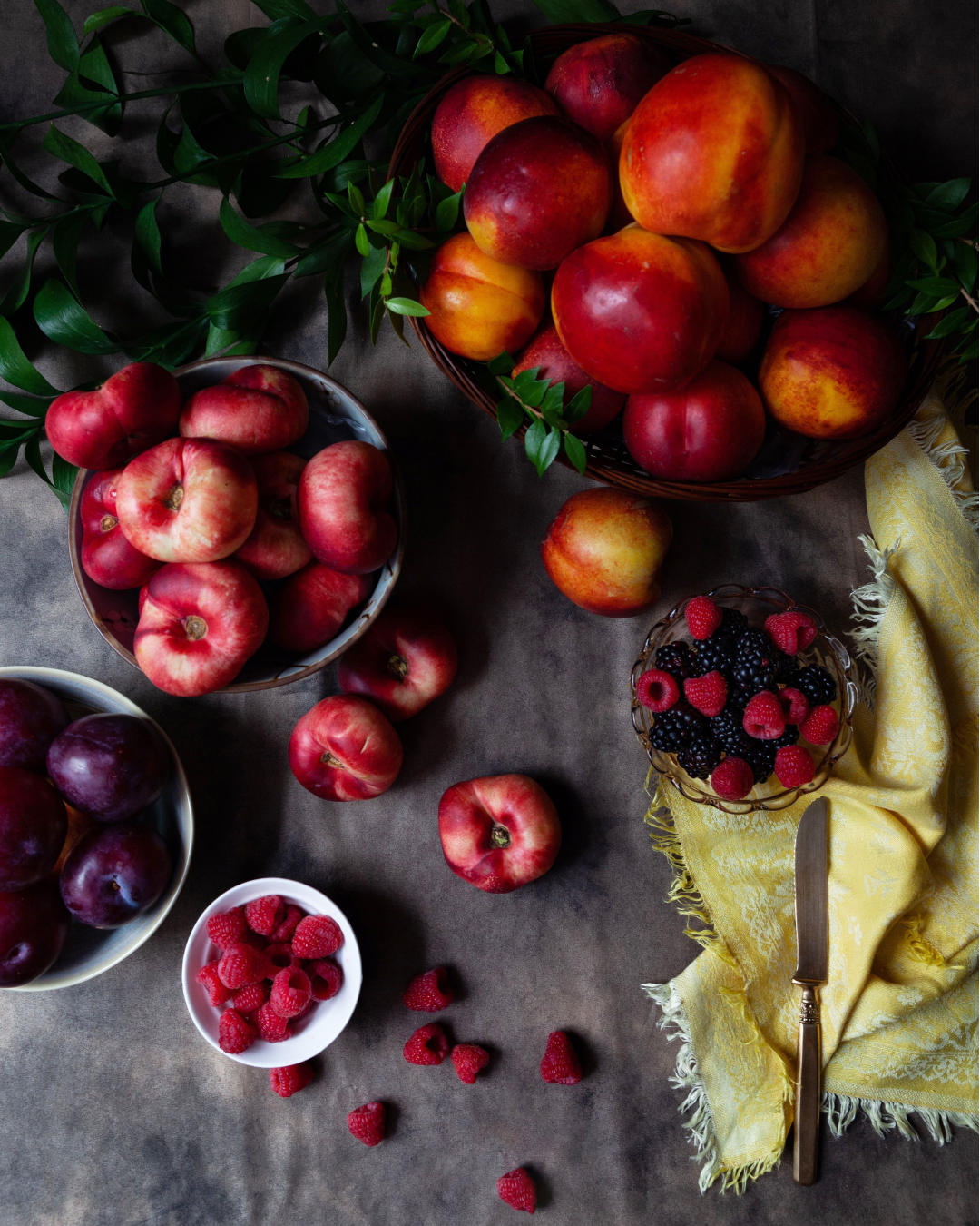 Ripe peaches, plums, raspberries, and blackberries arranged on a table among green branches