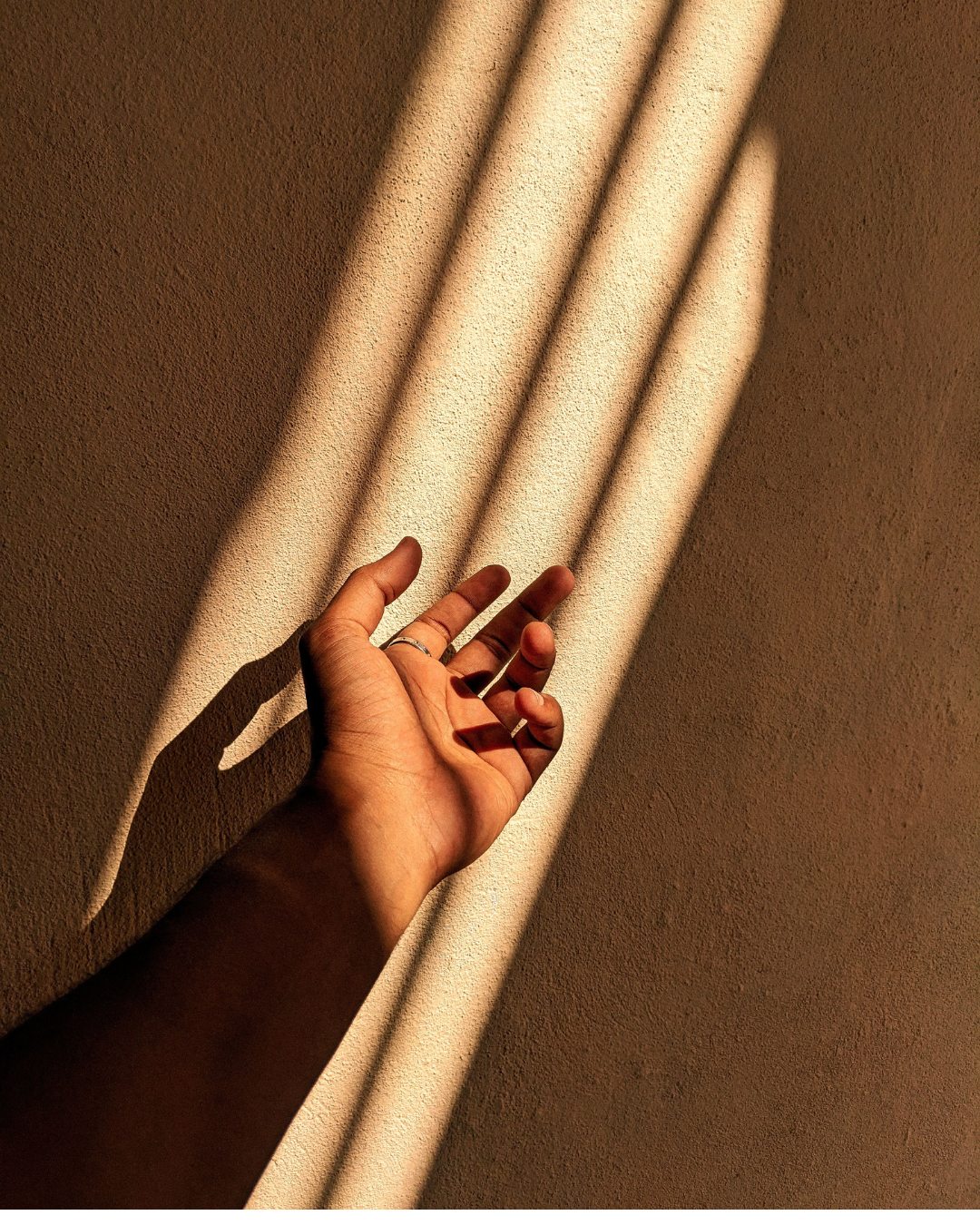 A hand reaching towards bands of light and shade on a warm, textured wall