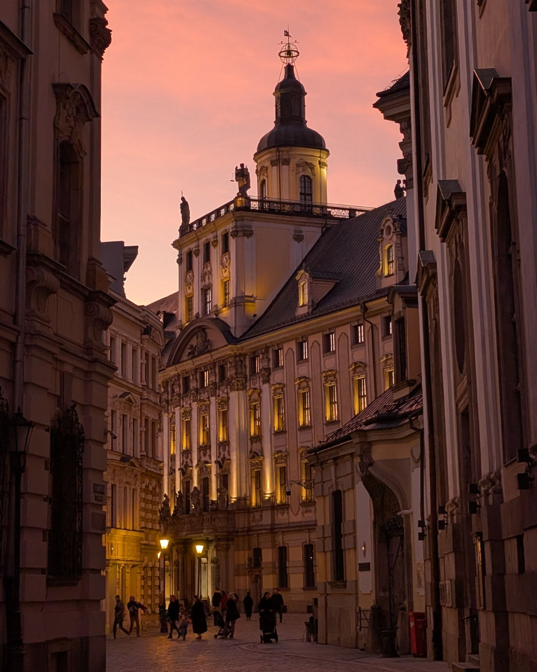 A historic city street at dusk with warm lights and a pink sky
